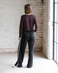 A person with short brown hair stands indoors, wearing a maroon long-sleeve top and Vassalli Asher Merino Pant - Black with a contrast side stripe. Natural light shines in from large windows on the right against a white brick wall.