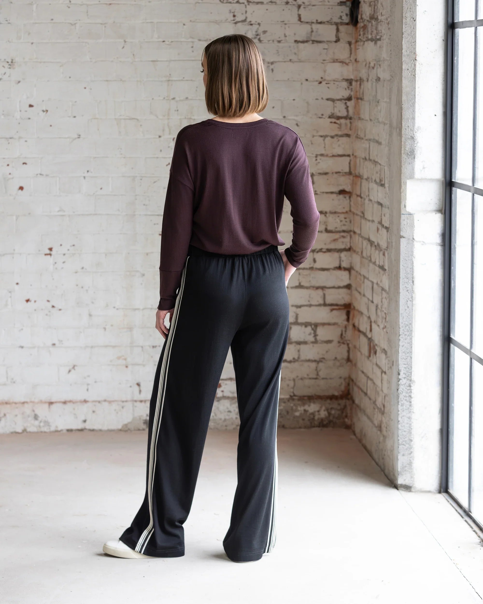 A person with short brown hair stands indoors, wearing a maroon long-sleeve top and Vassalli Asher Merino Pant - Black with a contrast side stripe. Natural light shines in from large windows on the right against a white brick wall.
