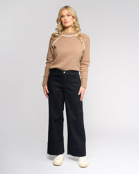 A woman with blonde hair faces forward, wearing Vassalli's Alley Jean in Black Denim, a light brown sweater with white trim, and white sneakers against a plain light gray background.