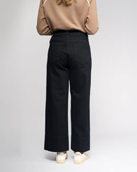 A person stands facing away from the camera, wearing a beige long-sleeve top, Vassalli Alley Jean - Black Denim, and white Veja sneakers against a plain light background.