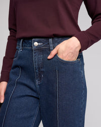A person is shown wearing a burgundy long-sleeve top and Vassalli Alley Jean in dark blue with white stitching, standing with one hand in their pocket. The image displays the torso and upper legs.