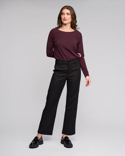 A woman stands against a light gray background, confidently posing in the Vassalli Neve Pant - Black, paired with a maroon long-sleeve top and black loafers.