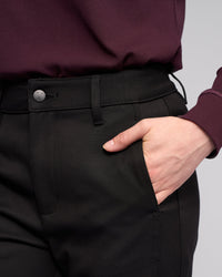 Model wears Vassalli’s Neve Pant in black with a burgundy long-sleeve top, right hand in pocket. The photo is cropped to highlight the waist and hand, without showing the model’s face.