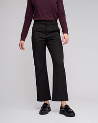A person models the Vassalli Neve Pant in black with a long-sleeve maroon top and shiny black loafers against a plain light gray background. Their head is not shown in the image.