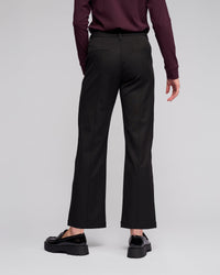A person stands facing away against a light gray background, wearing Vassalli's Neve Pant in black, a long-sleeved maroon top, and black loafers.