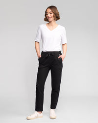 A person with short brown hair smiles against a plain background, wearing a white V-neck T-shirt, Vassalli Sunday Pant - Black with an elasticated waistband and slim leg, and white sneakers, hands in pockets.