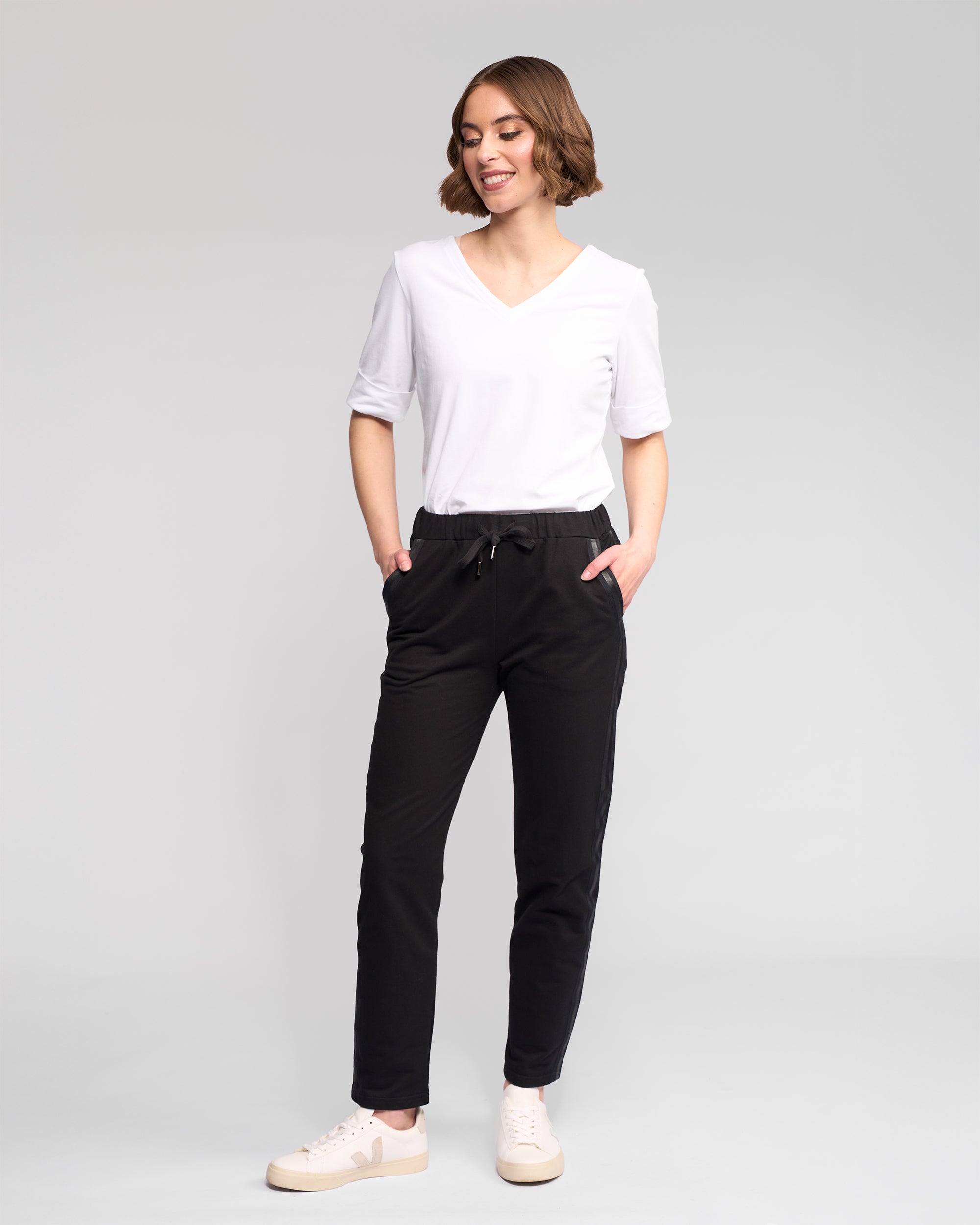 A person with short brown hair smiles against a plain background, wearing a white V-neck T-shirt, Vassalli Sunday Pant - Black with an elasticated waistband and slim leg, and white sneakers, hands in pockets.