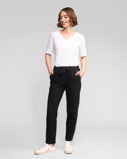 A person with short brown hair smiles against a plain background, wearing a white V-neck T-shirt, Vassalli Sunday Pant - Black with an elasticated waistband and slim leg, and white sneakers, hands in pockets.