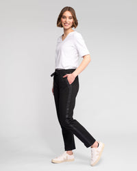 A woman with short brown hair smiles with one hand in her pocket, wearing a white top and Vassalli Sunday Pant - Black, featuring a slim leg, elasticated waistband, and side stripe, against a plain light background.