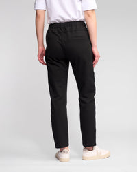 A person stands facing away, wearing a white t-shirt and Vassalli Sunday Pant - Black joggers with a slim leg and elastic waistband, paired with off-white Veja sneakers. The background is plain and light-colored.