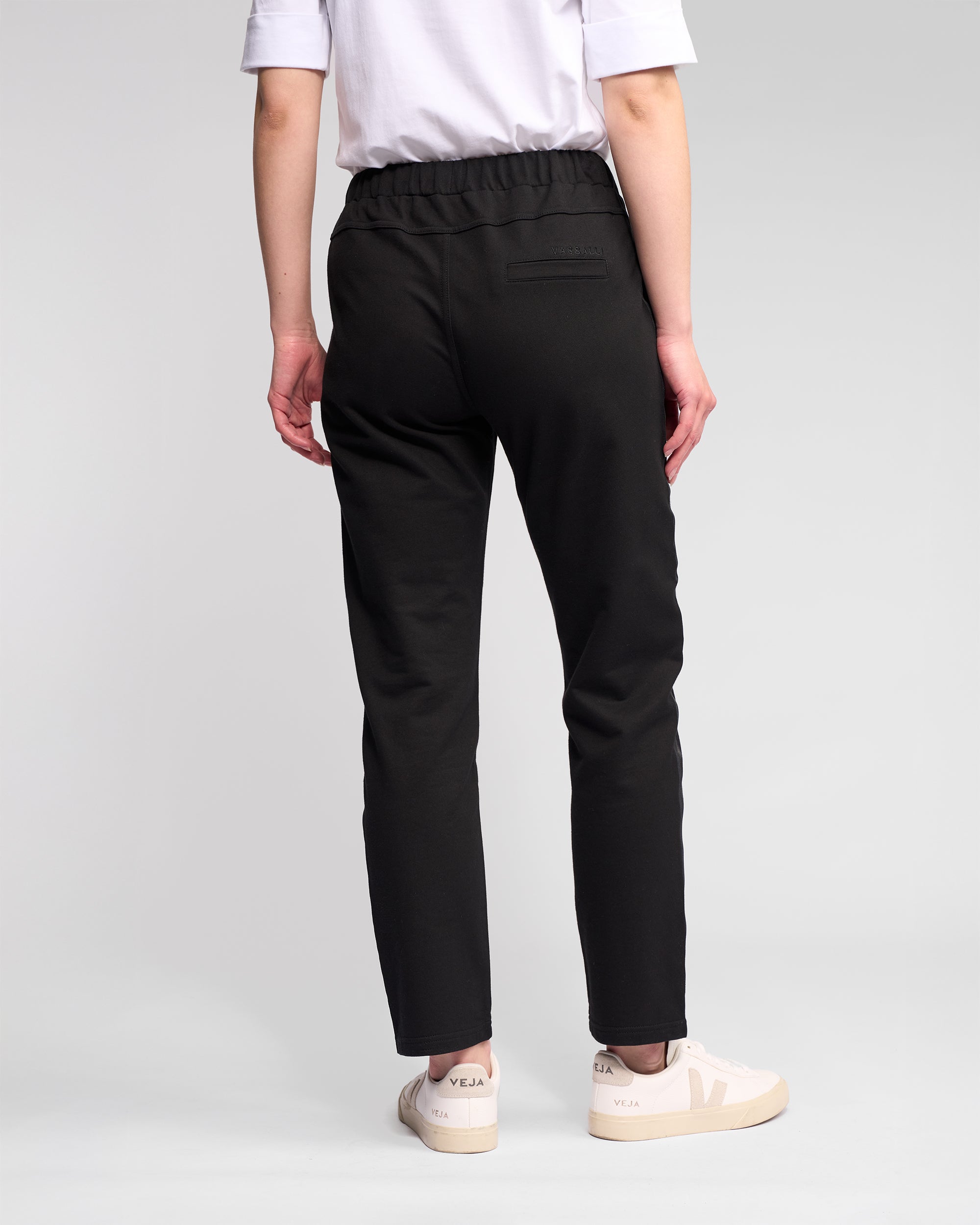 A person stands facing away, wearing a white t-shirt and Vassalli Sunday Pant - Black joggers with a slim leg and elastic waistband, paired with off-white Veja sneakers. The background is plain and light-colored.