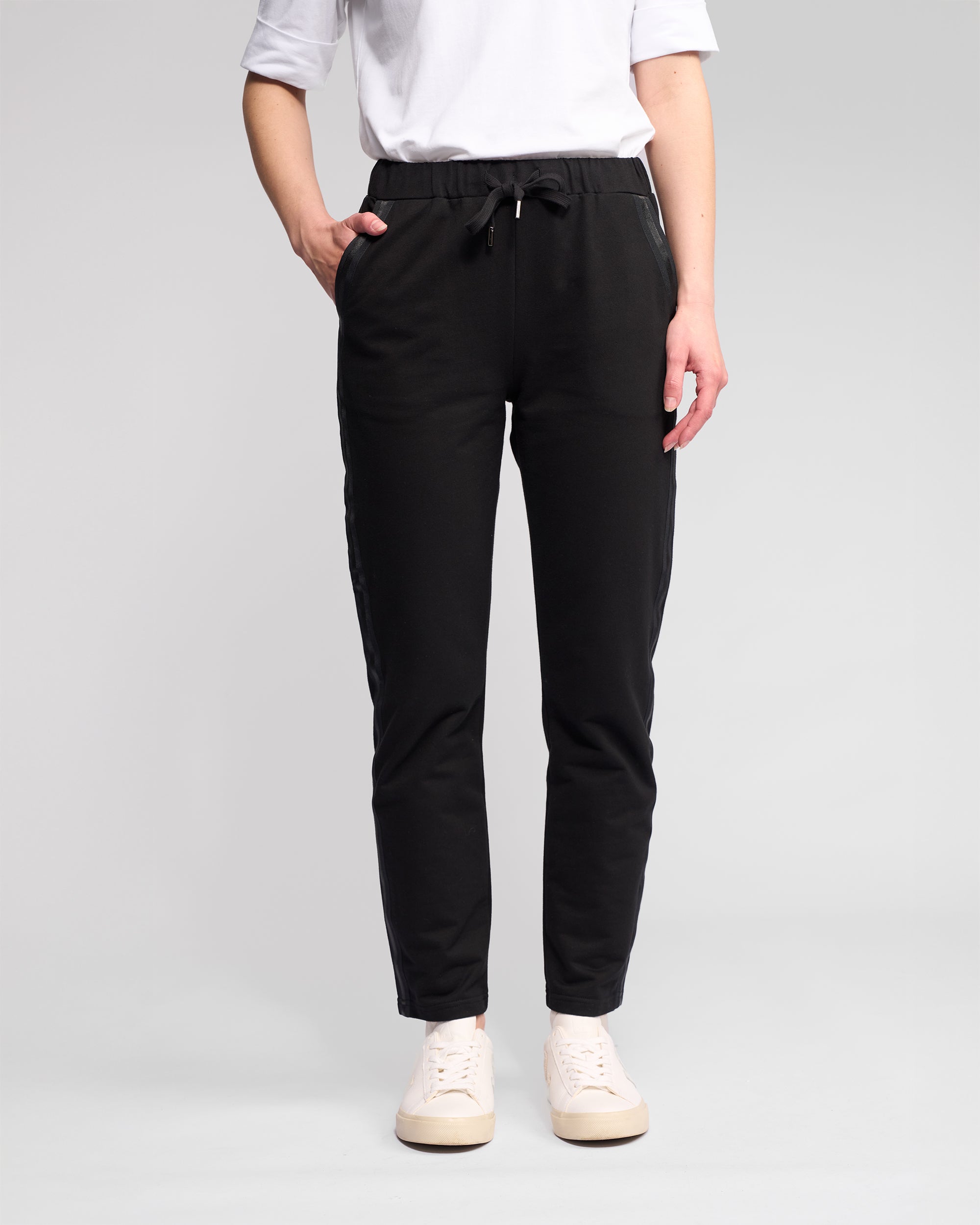 Wearing a white T-shirt, white sneakers, and Vassalli Sunday Pant - Black with a slim leg and elasticated waistband, a person stands with one hand in their pocket against a plain light background.