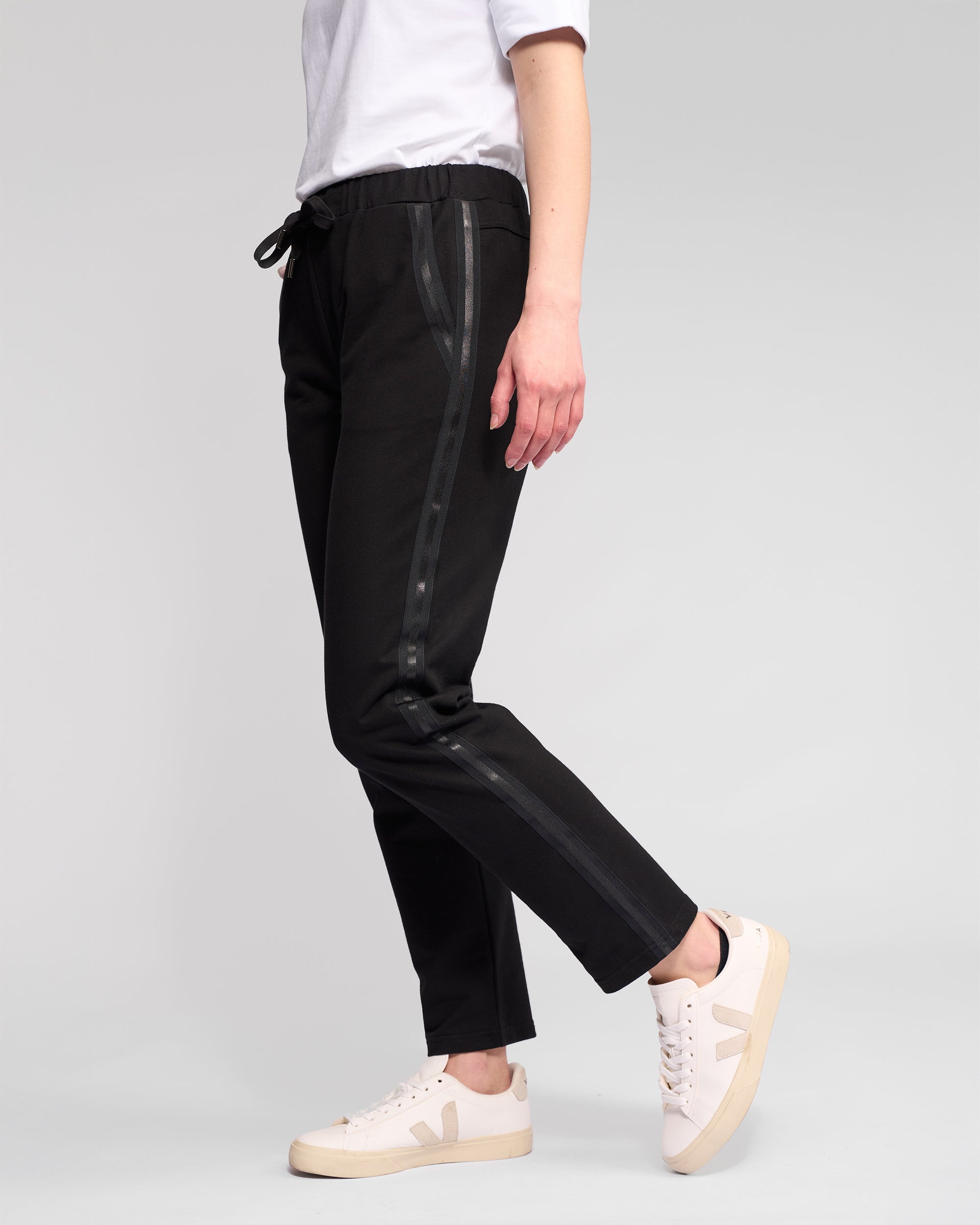 A person stands against a light gray background wearing Vassalli's Sunday Pant in black, featuring a shiny side stripe, slim leg fit, and elasticated waistband. Off-white sneakers with beige soles and a V logo complete the look. Head is out of frame.