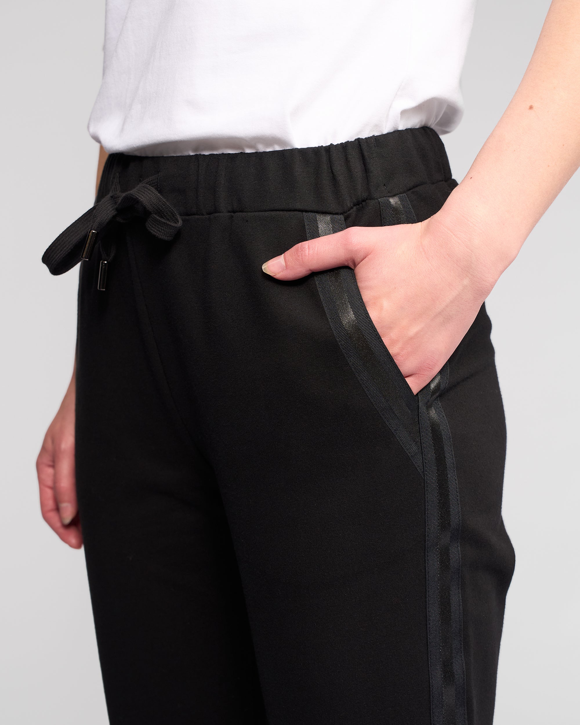 Someone is shown from the waist down wearing Vassalli's Sunday Pant in Black—a casual jogger with an elasticated waistband and slim leg—paired with a white shirt, one hand in their pocket, against a plain background.