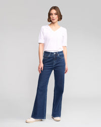 A person with short, wavy brown hair and a neutral expression stands against a plain light gray background, wearing Vassalli's Wren Jean in Dark Blue, a white short-sleeve V-neck T-shirt, and white sneakers.