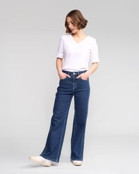 A person stands with hands in pockets, looking down, wearing a white V-neck top, Vassalli Wren Jean in dark blue, and white sneakers against a plain light gray background.