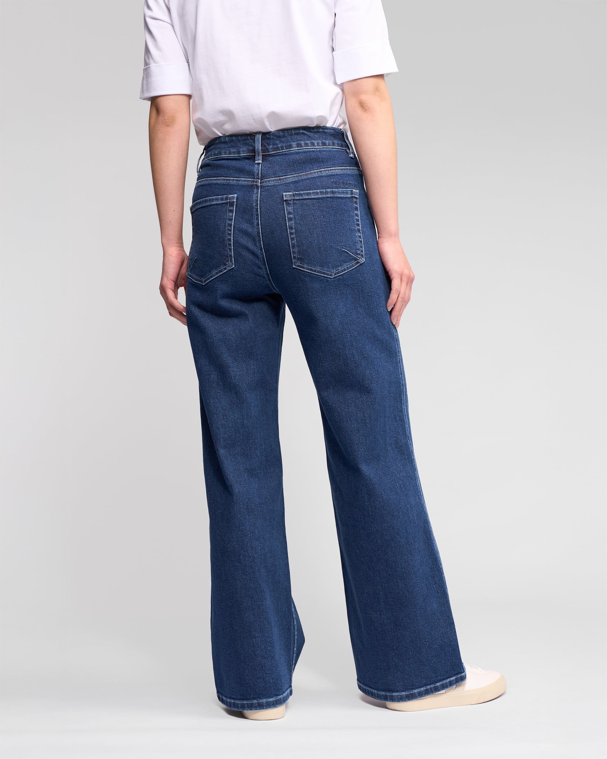 A person stands facing away, wearing a white short-sleeve shirt, Vassalli’s Wren Jean in dark blue—high-waisted, full-length, wide-leg—paired with white sneakers against a plain light background.