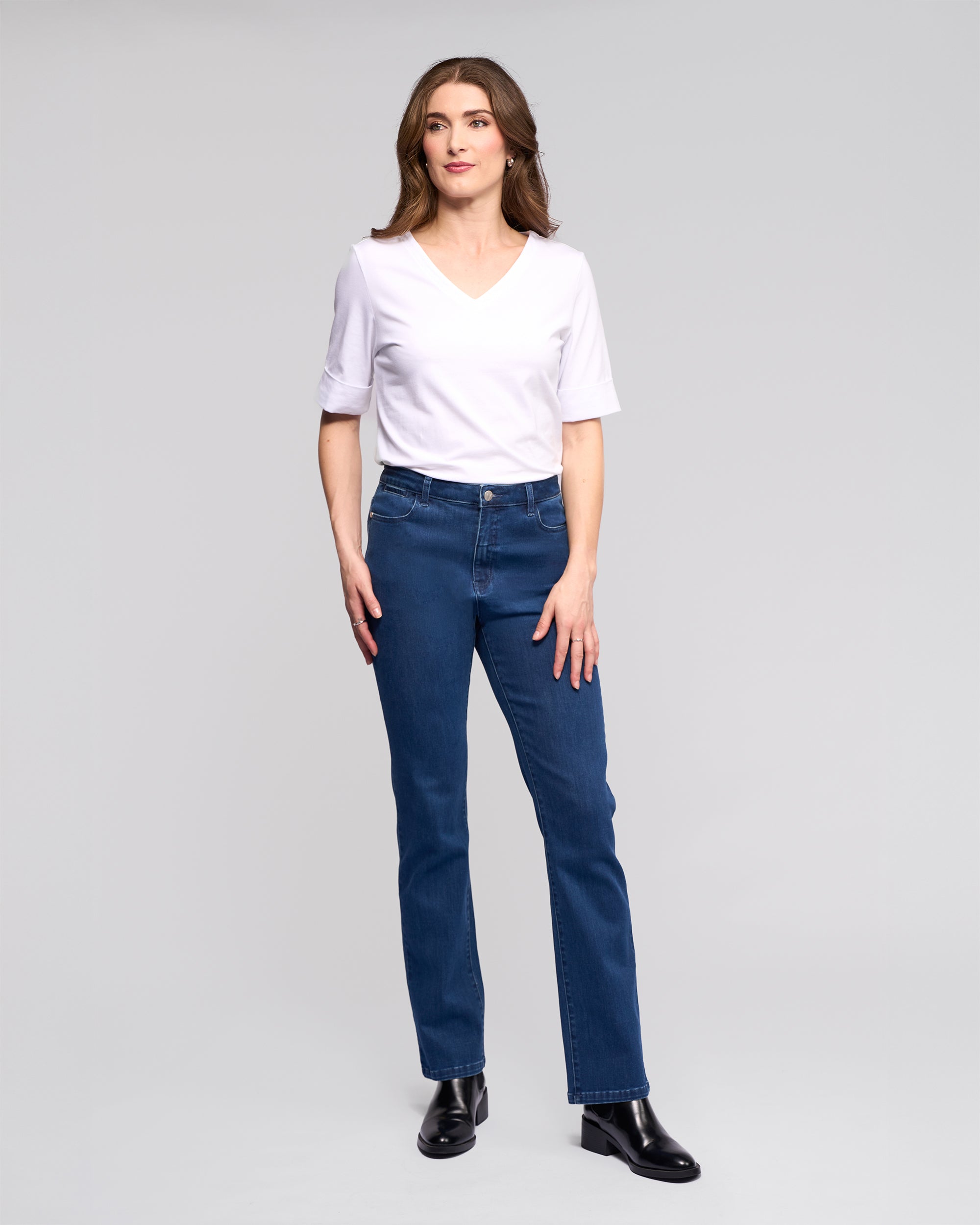 A woman stands against a light gray background, wearing Vassalli's Radio Jean in blue denim, a white V-neck T-shirt, and black ankle boots. She has long brown hair and gazes slightly to the side with a neutral expression.