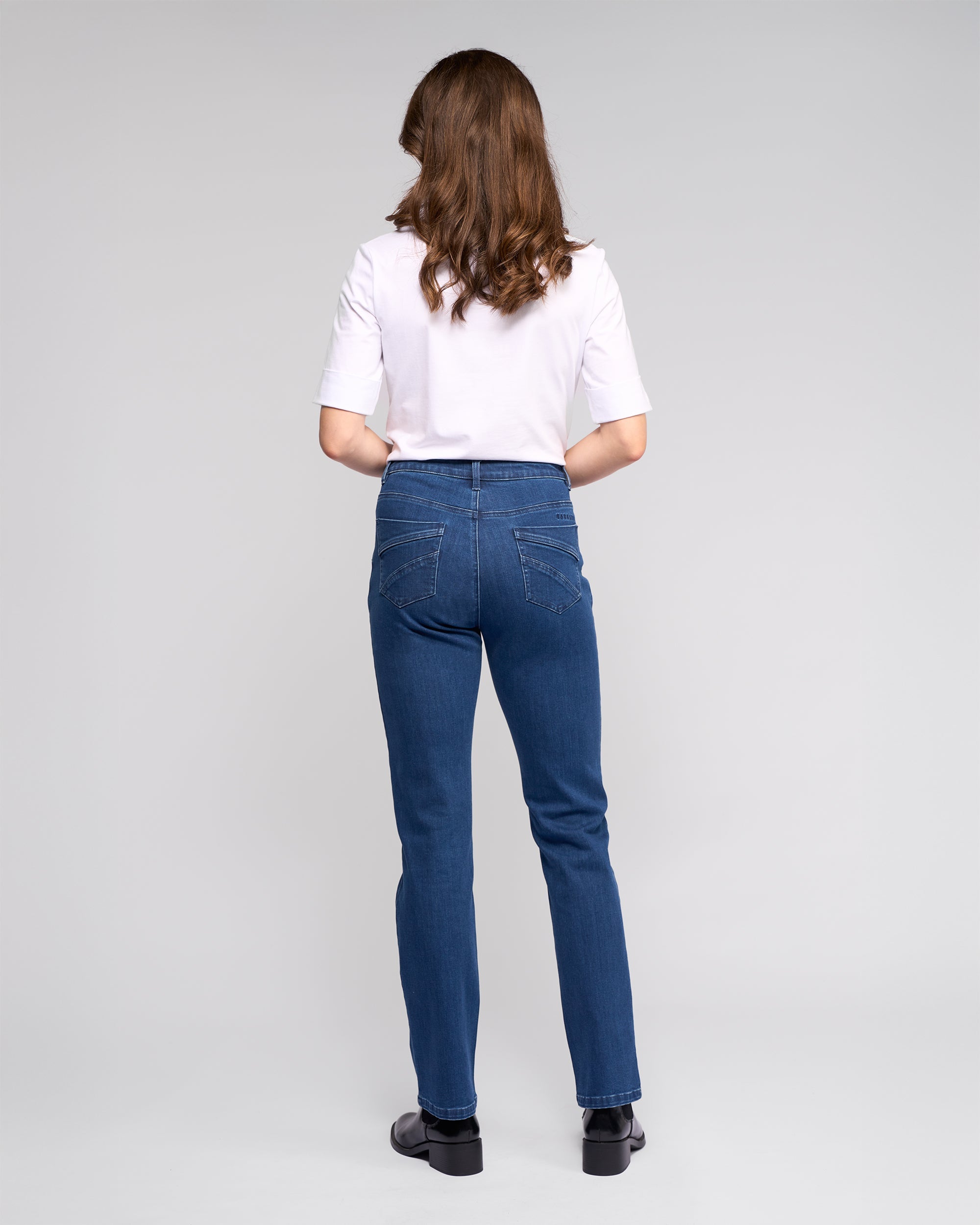 A woman with wavy brown hair faces away, wearing a white short-sleeve shirt, black shoes, and Vassalli's Radio Jean in Blue Denim against a plain light gray background.