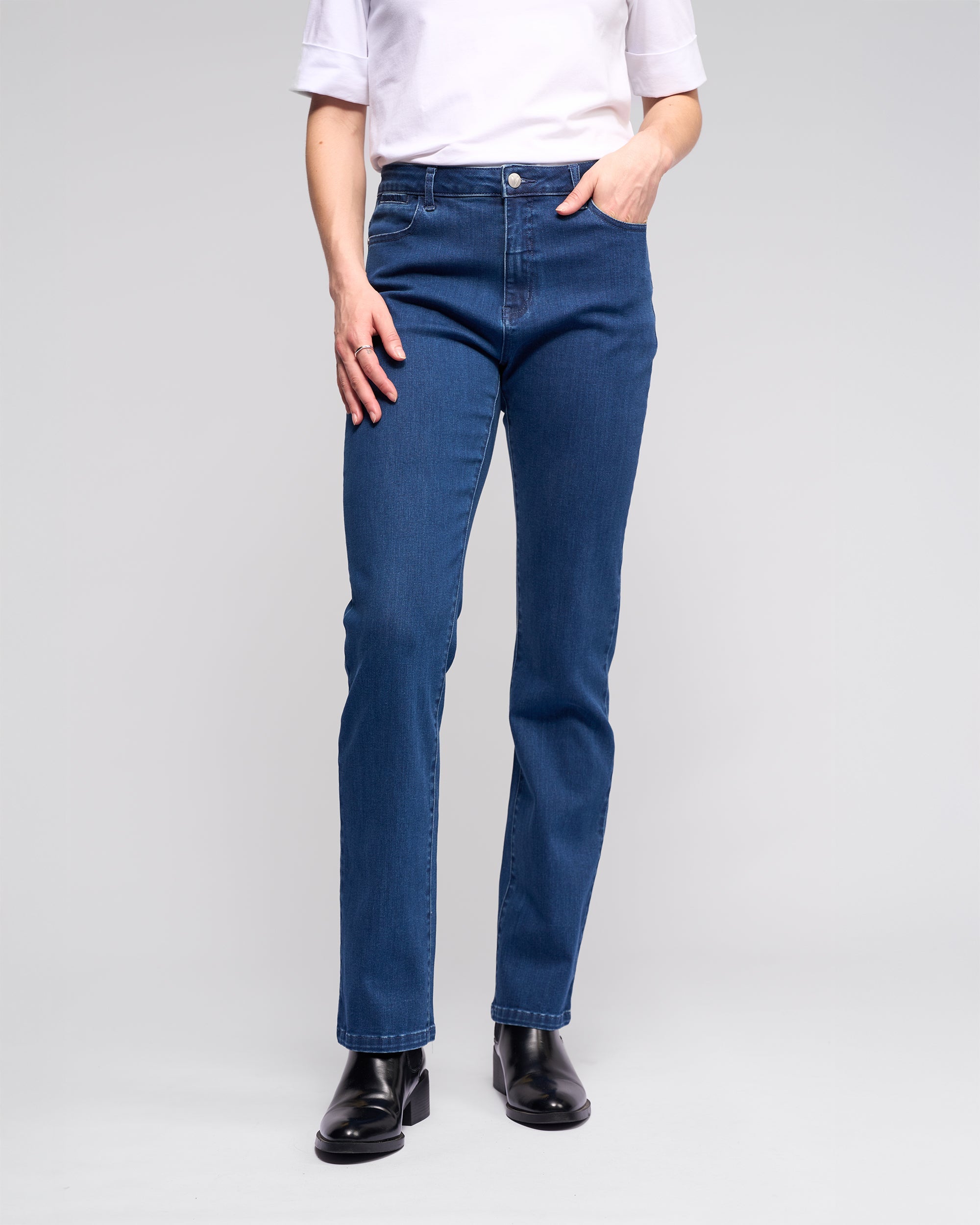 A person wears a white short-sleeve top, Vassalli Radio Jean - Blue Denim, and black ankle boots while standing against a plain light background.