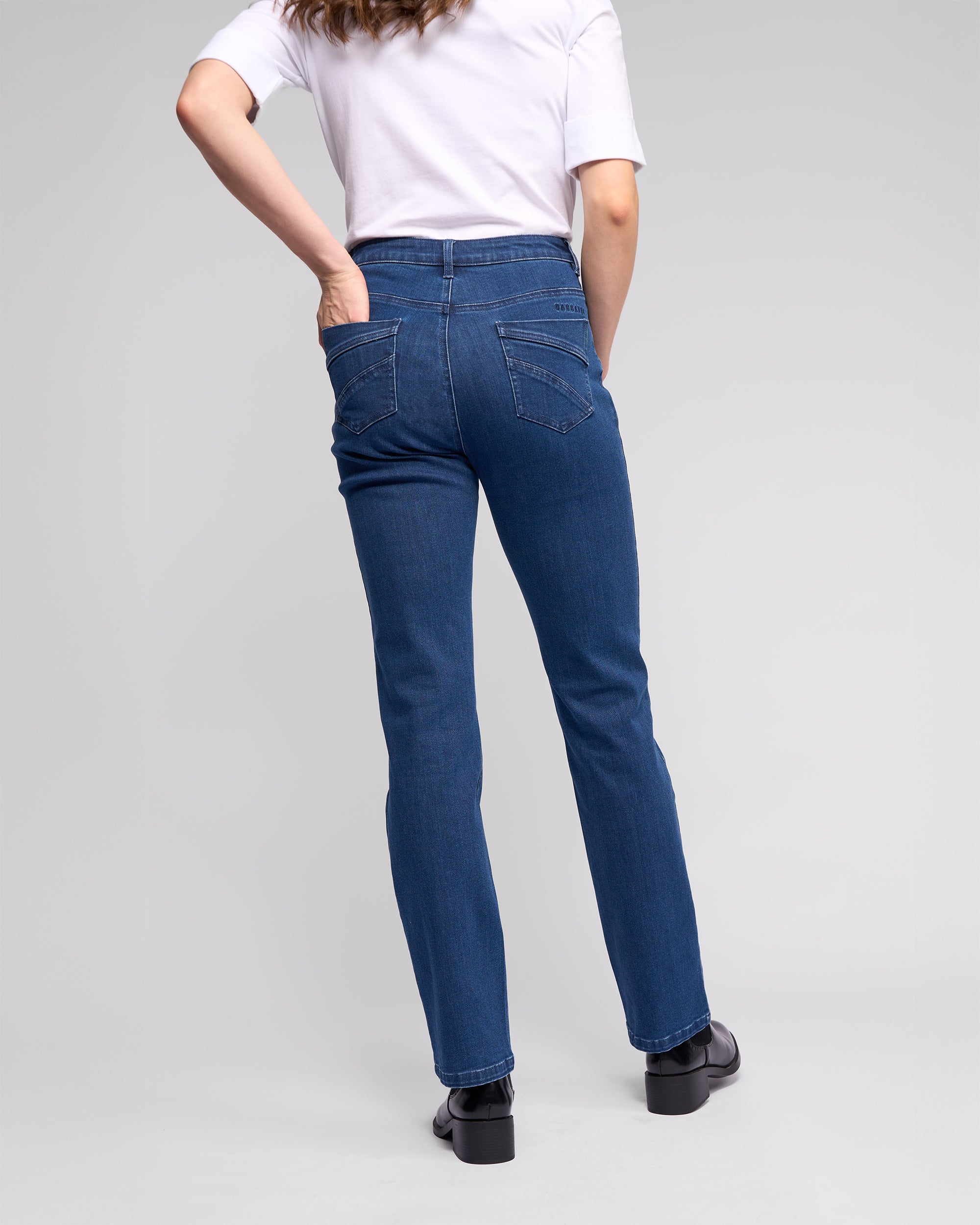 A person stands facing away with one hand on their hip, wearing a white short-sleeve shirt, black shoes, and Vassalli's Radio Jean in Blue Denim against a plain light gray background.