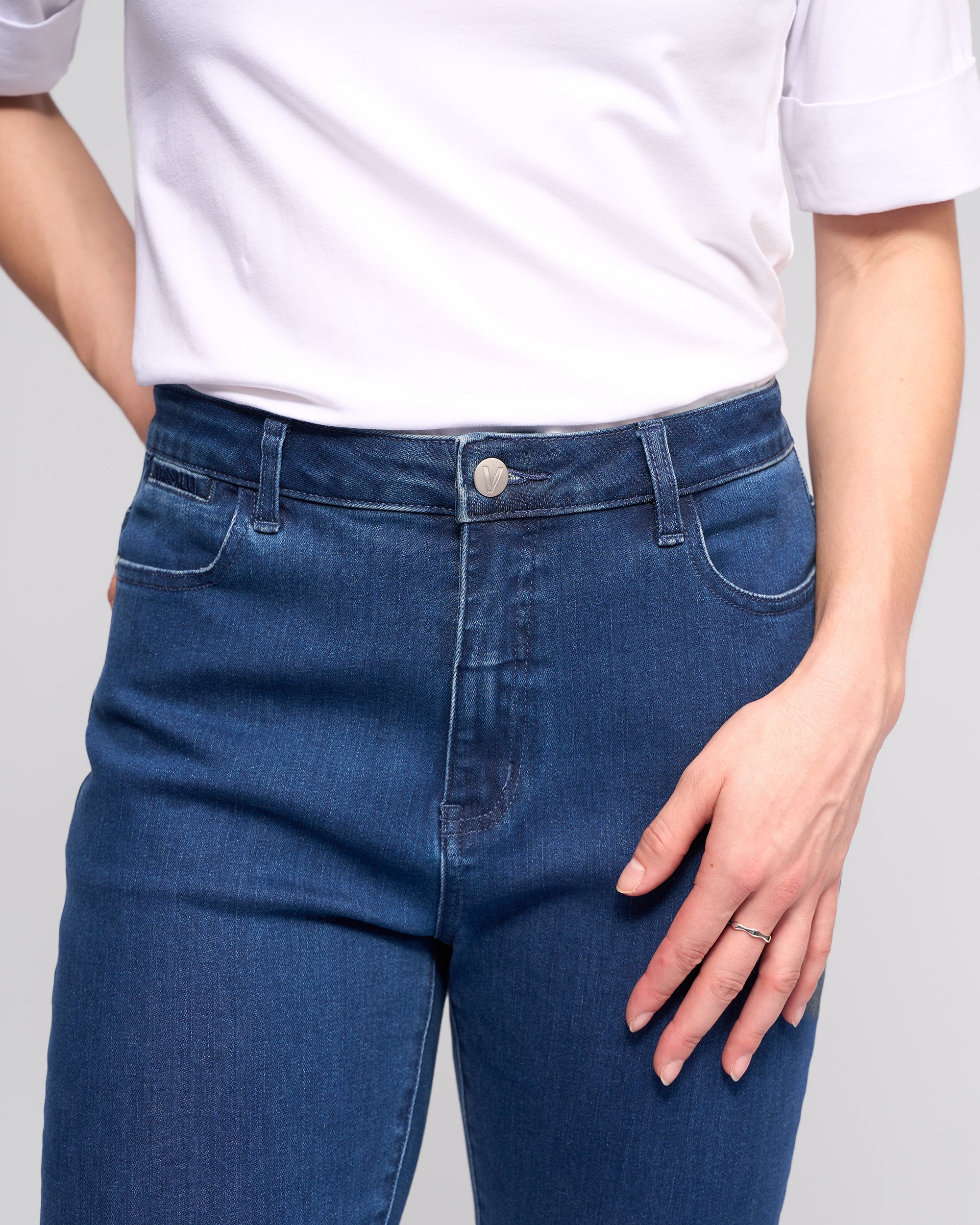 Wearing a white short-sleeve shirt and Vassalli Radio Jean - Blue Denim, a person poses with one hand on their hip and the other relaxed by their side, displaying a simple ring.