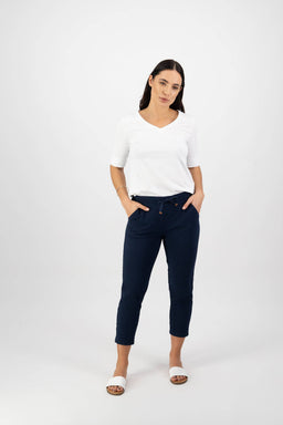 A woman with long dark hair wears a white V-neck top, Vassalli Denver 7/8 Pant in Ink, and white slide sandals, standing against a plain white background with her hands in her pockets.