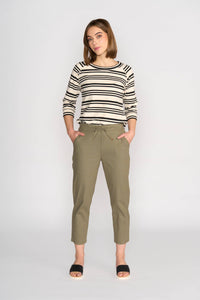 A woman stands against a plain background, wearing a black and white striped long-sleeve top, Vassalli Henley 7/8 Pant - Army, and black open-toe sandals, with her hands in her pockets.