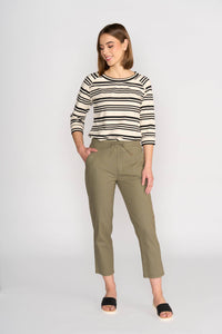 A woman smiles and looks to the side, wearing a black and white striped long-sleeve top, Vassalli's Henley 7/8 Pant in Army, and black slide sandals, against a plain light background.
