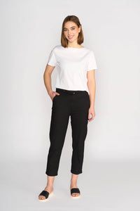 A woman stands against a white background, smiling. She wears a white short-sleeve tee and Vassalli Henley 7/8 Pant - Black, featuring an elastic waistband and slim leg, paired with black slip-on sandals. One hand is in her pocket.