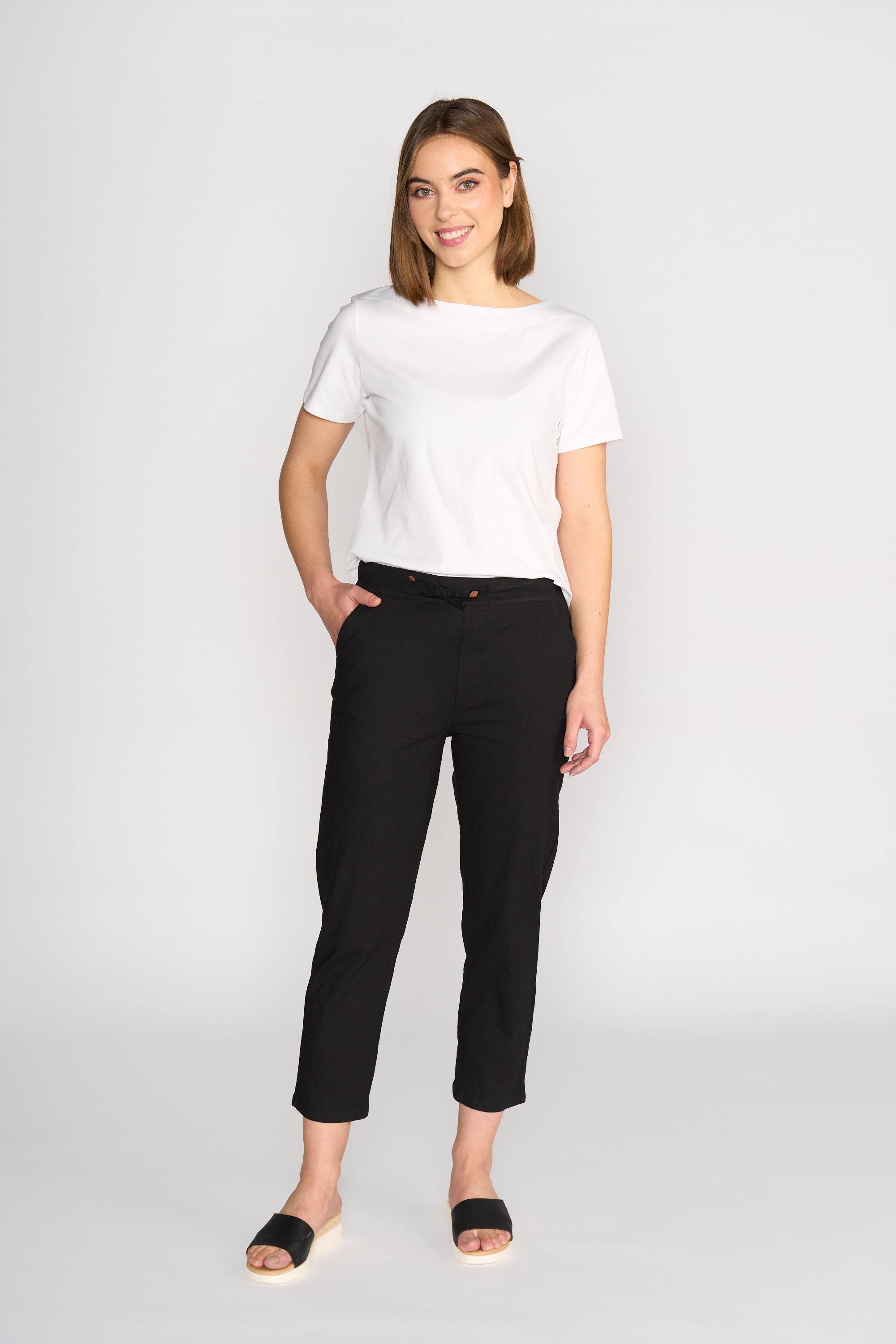 A woman stands against a white background, smiling. She wears a white short-sleeve tee and Vassalli Henley 7/8 Pant - Black, featuring an elastic waistband and slim leg, paired with black slip-on sandals. One hand is in her pocket.