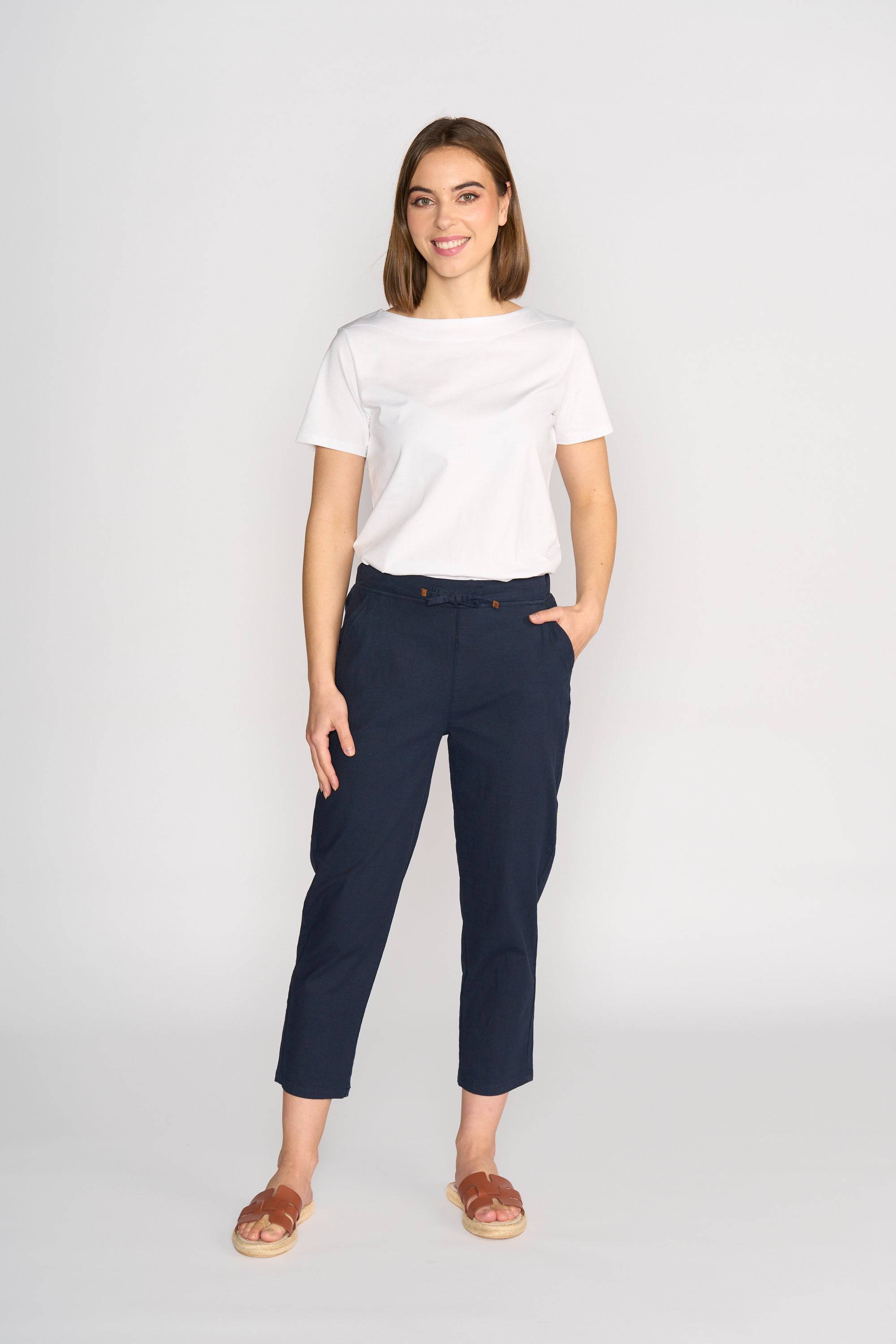 A woman smiles against a plain background, wearing a white short-sleeve tee, brown sandals, and the Vassalli Henley 7/8 Pant in Ink with an elastic waistband and slim-leg fit; one hand rests in her pocket.