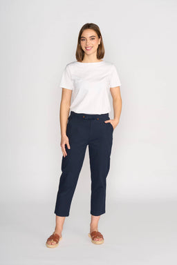 A woman smiles against a plain background, wearing a white short-sleeve tee, brown sandals, and the Vassalli Henley 7/8 Pant in Ink with an elastic waistband and slim-leg fit; one hand rests in her pocket.