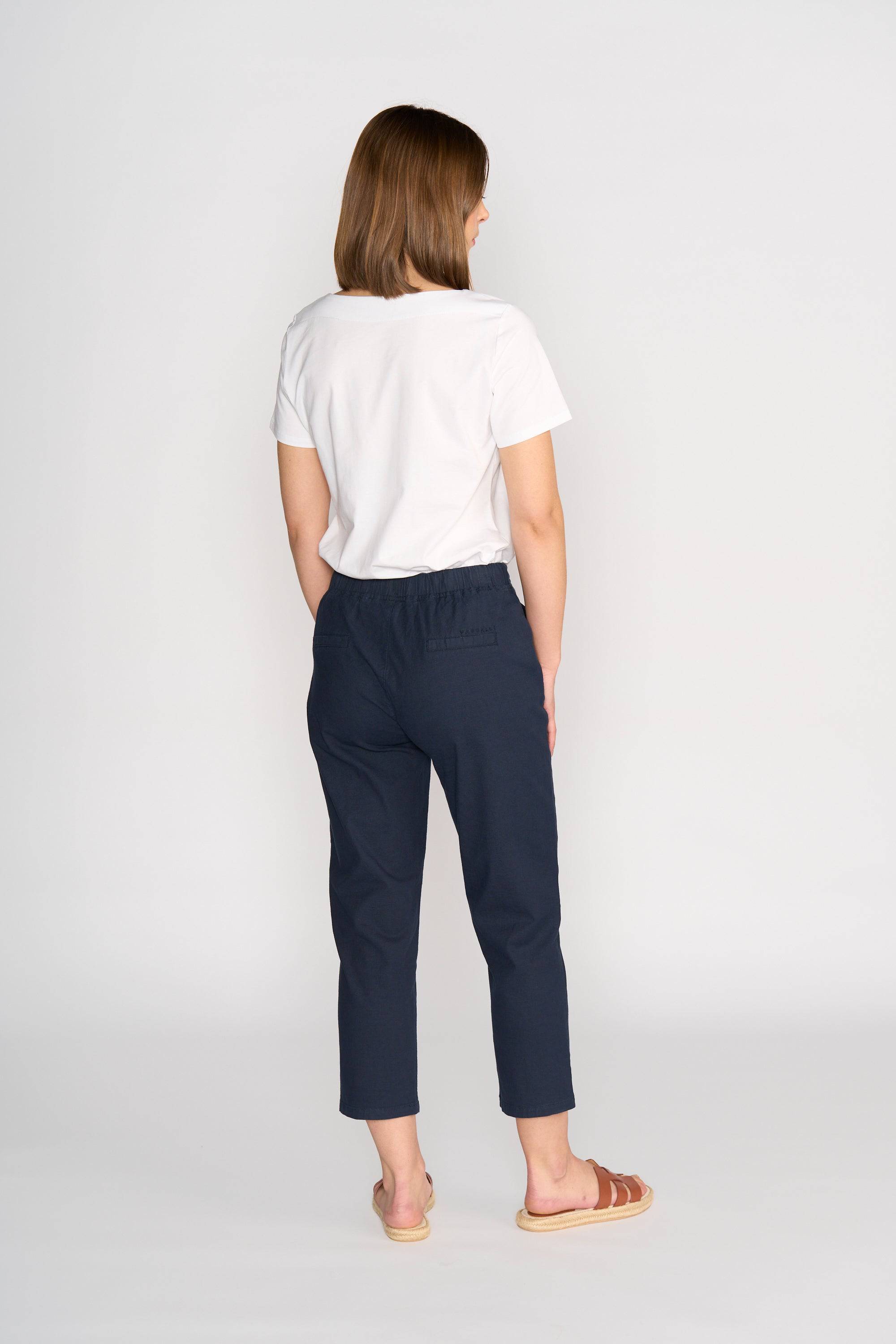 A person with shoulder-length brown hair wears a white short-sleeve t-shirt, Vassalli Henley 7/8 Pant – Ink, and brown sandals, standing facing away from the camera against a plain light background.