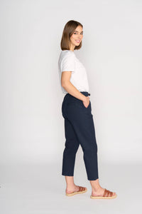 A woman with straight brown hair stands in profile, smiling with her hands in her pockets, wearing a white t-shirt and the Vassalli Henley 7/8 Pant in Ink against a plain light background.