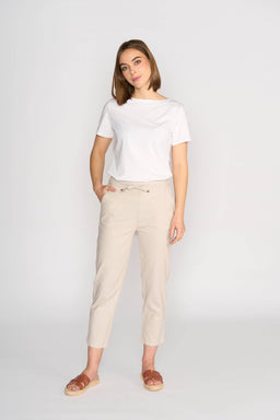 A woman stands against a plain white background wearing Vassalli’s Henley 7/8 Pant in Stone, a pair of beige slim-leg pants with an elastic waistband, paired with a white t-shirt and brown sandals. She poses with one hand in her pocket.