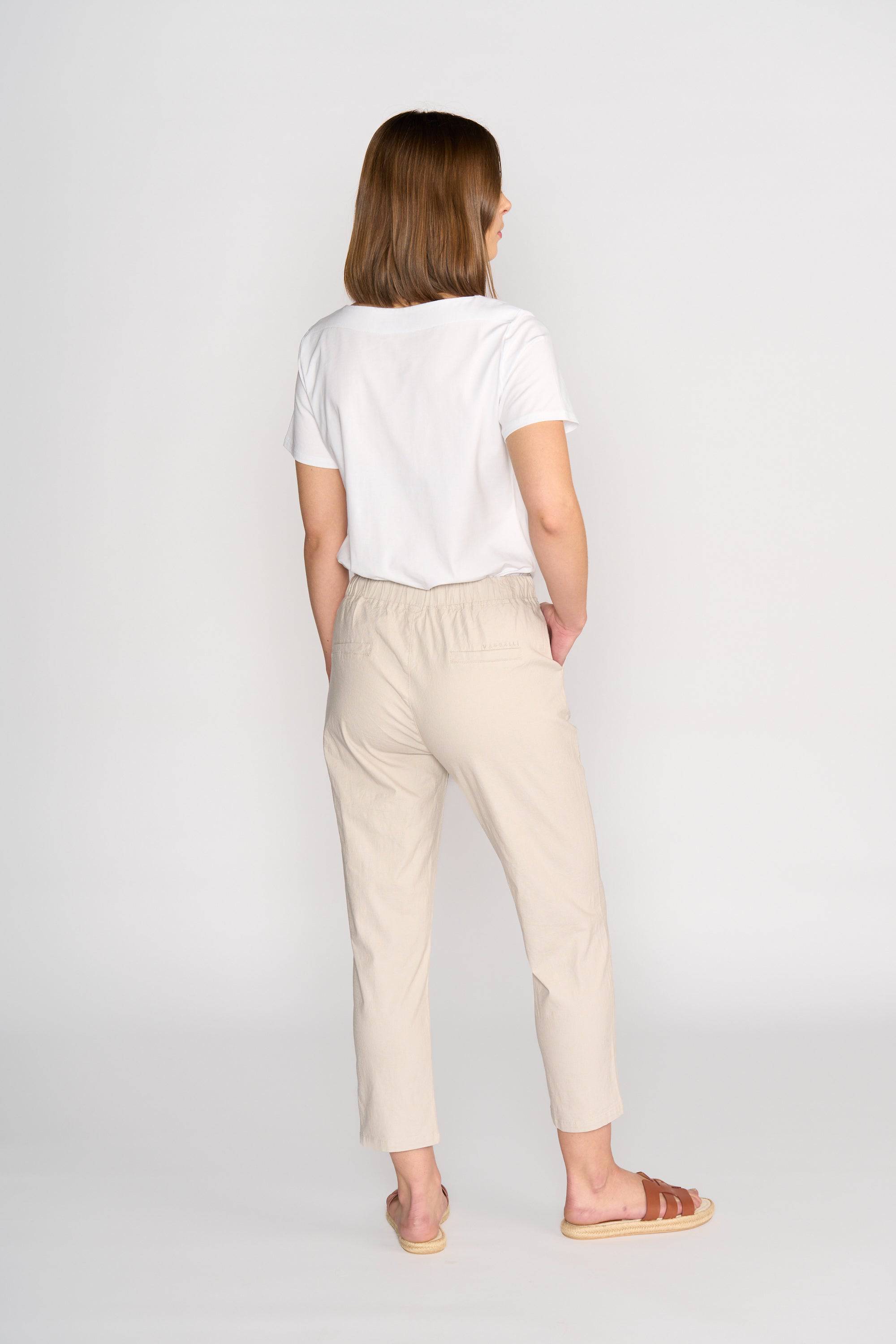 A woman faces away from the camera, wearing a white T-shirt and Vassalli Henley 7/8 Pant in Stone with an elastic waistband, paired with brown sandals, set against a plain light background.