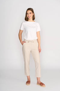 A woman with chin-length hair smiles against a light background, wearing a white t-shirt, brown sandals, and Vassalli's Henley 7/8 Pant in Stone—a slim-leg, light beige style with an elastic waistband and drawstring. Her left hand is in her pocket.