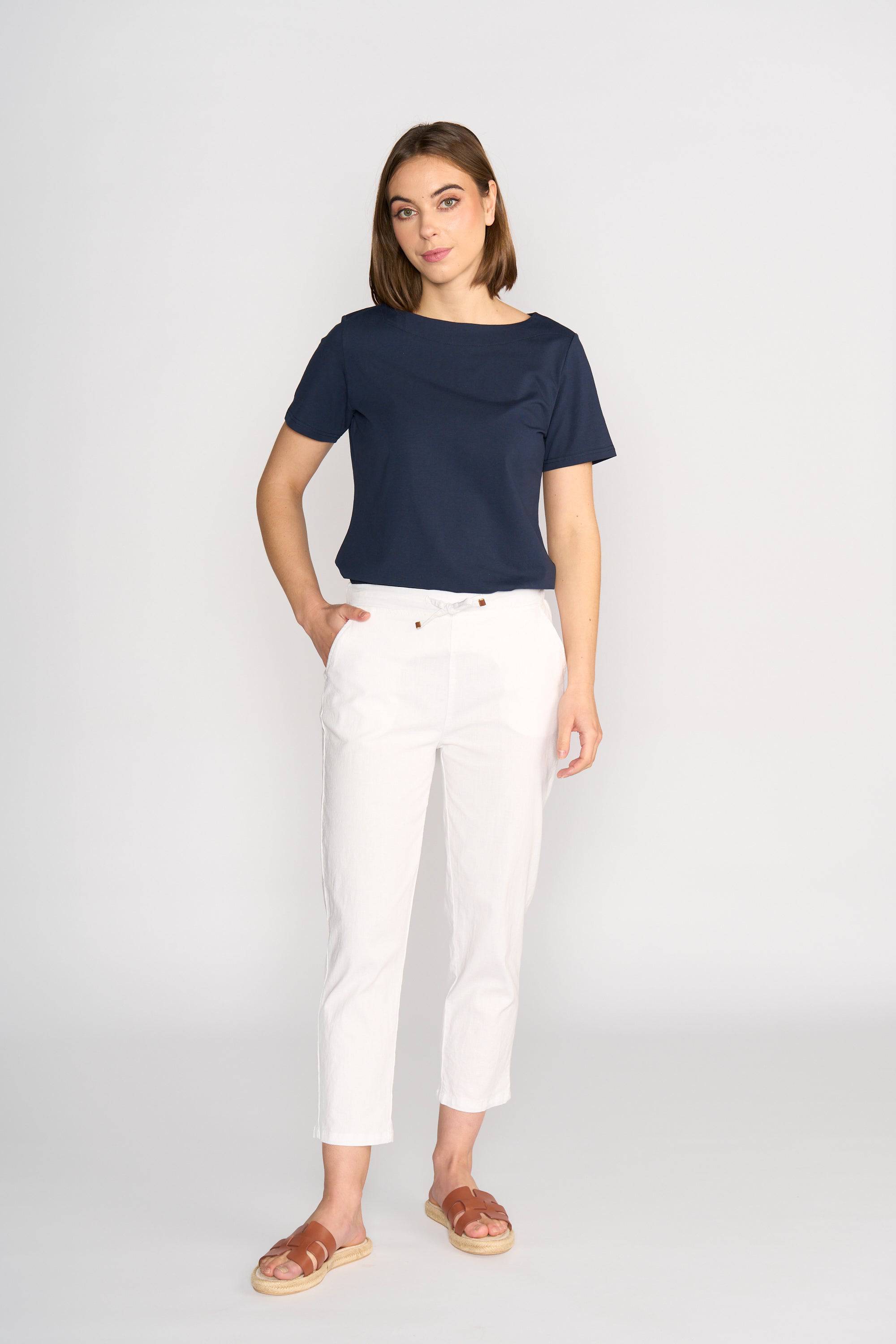 A woman with shoulder-length brown hair stands against a plain light background, wearing a navy blue short-sleeve top, brown sandals, and Vassalli's Henley 7/8 Pant in white with an elastic waistband. One hand is in her pocket.