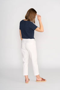 A woman, facing away, wears the Vassalli Henley 7/8 Pant in white with a navy t-shirt and brown sandals. Her left arm is raised near her head and her chin turns slightly right. The background is plain and light-colored.