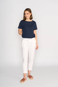 A woman smiles while looking down, wearing the Vassalli Henley 7/8 Pant in white, paired with a navy short-sleeve top and brown sandals. She stands against a light background with one hand in her pocket.