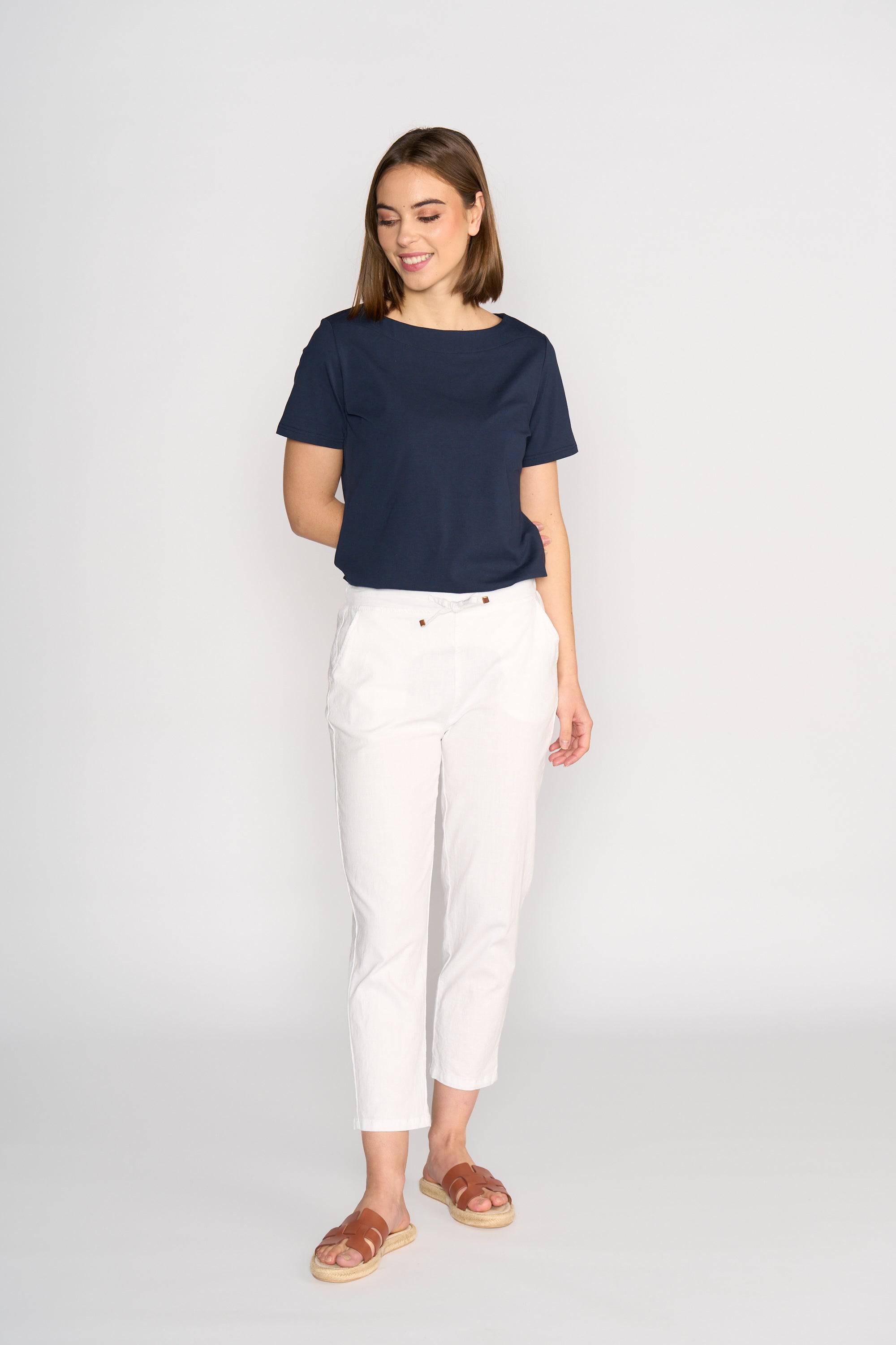 A woman smiles while looking down, wearing the Vassalli Henley 7/8 Pant in white, paired with a navy short-sleeve top and brown sandals. She stands against a light background with one hand in her pocket.
