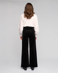 A woman with wavy brown hair, wearing a white blouse and Vassalli Empire Cord Pant in black, stands facing away from the camera against a plain light gray background.