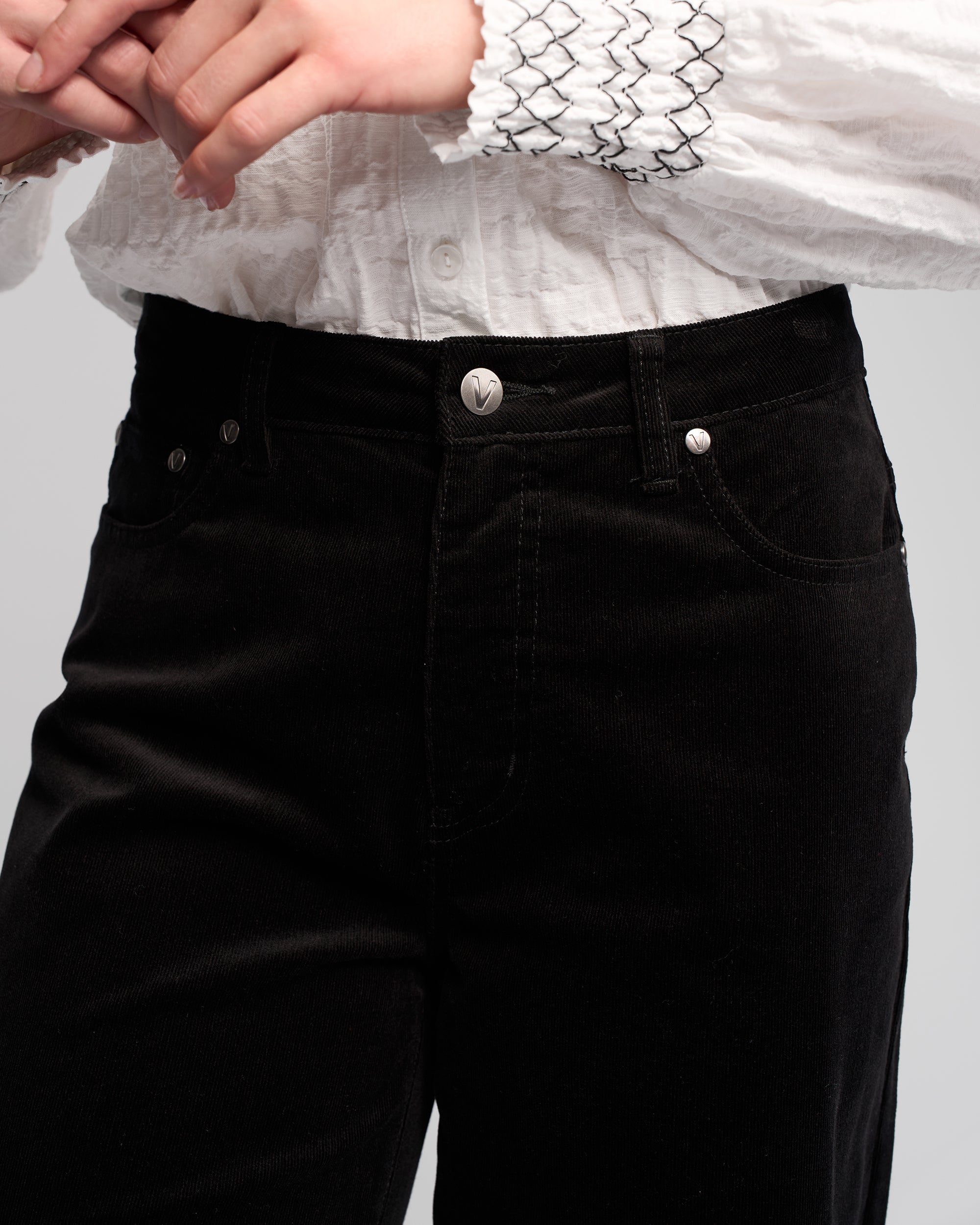 A person is shown from waist to mid-thigh wearing Vassalli's Empire Cord Pant in black, paired with a textured white blouse featuring smocked cuffs, standing with their hands clasped together.