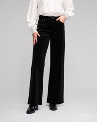 Model wears the Vassalli Empire Cord Pant in Black, paired with a textured white long-sleeve blouse with decorative cuffs and black heeled boots. Photo is cropped at the shoulders, set against a neutral background.