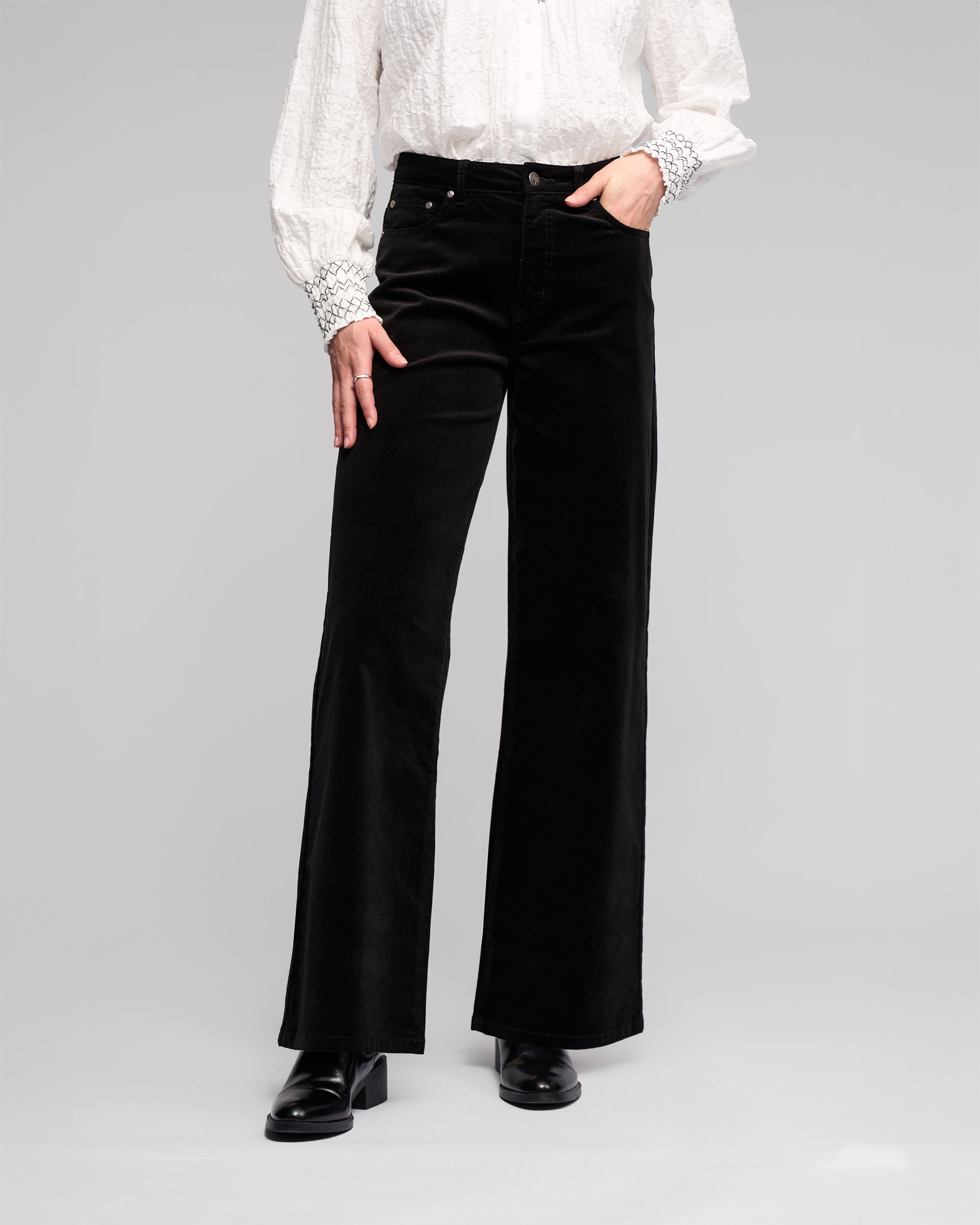 Model wears the Vassalli Empire Cord Pant in Black, paired with a textured white long-sleeve blouse with decorative cuffs and black heeled boots. Photo is cropped at the shoulders, set against a neutral background.
