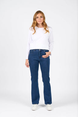 A woman with long blonde hair wears the Vassalli Theory Jean in blue denim (without fade), a white long-sleeve shirt, and white shoes. She stands facing forward with one hand in her pocket against a plain white background.