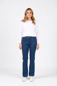 Woman wearing a white long-sleeve cropped top and Vassalli Theory Jean in blue denim (without fade) with white trainers, standing against a light grey background.