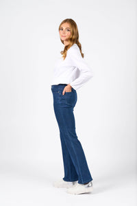 A woman with long light brown hair, in a white long-sleeve shirt, Vassalli Theory Jean - Blue Denim (Without Fade), and white sneakers stands sideways with one hand in her pocket, looking at the camera against a plain white background.