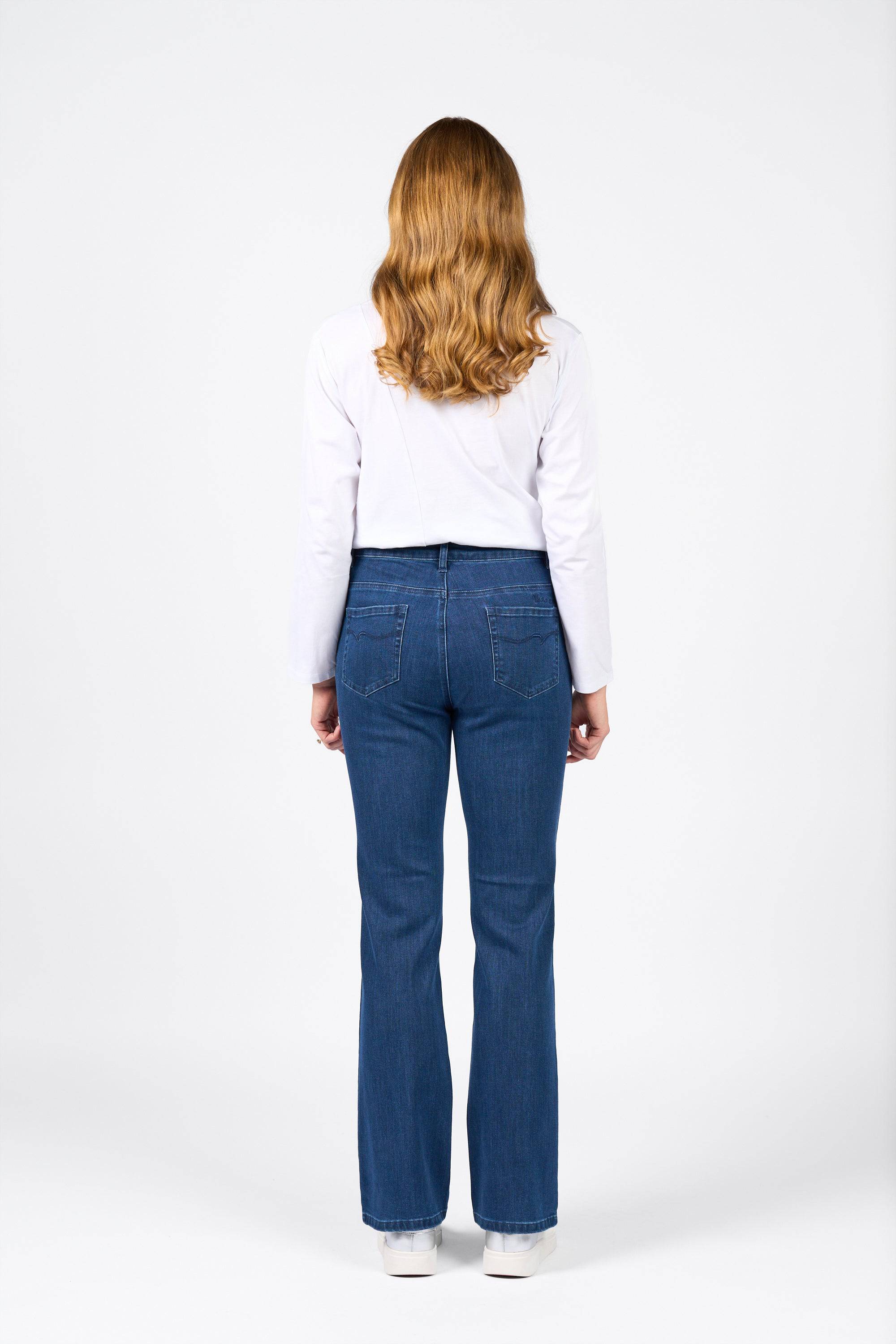 A person with long, wavy blond hair is standing with their back to the camera, wearing a white long-sleeve shirt, Vassalli Theory Jean - Blue Denim (Without Fade), and white shoes against a plain white background.